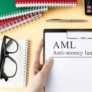 Dłoń trzyma dokument z tekstem AML Anti-money laundering, obok okulary, długopis, kolorowe kołonotatniki.